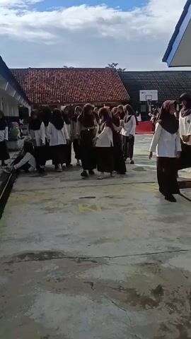A group of adolescents are walking through an outdoor area. Most are wearing school uniforms consisting of white shirts and black pants or skirts, and brown hijabs. Some students are adjusting their headscarves, and one is eating. The background includes parts of school buildings with a red-tiled roof and a basketball hoop. The ground they are walking on is concrete with yellow painted letters, suggesting a designated area or path. The sky is blue with scattered clouds, indicating it's daytime.