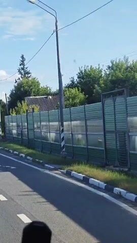 A video shows a car driving by a road. There is a crosswalk sign and a bus stop shelter. A green and transparent sound barrier is visible along the side of the road. Trees and houses can be seen in the background. The sky is blue with some clouds. The road has white lines and a curb.