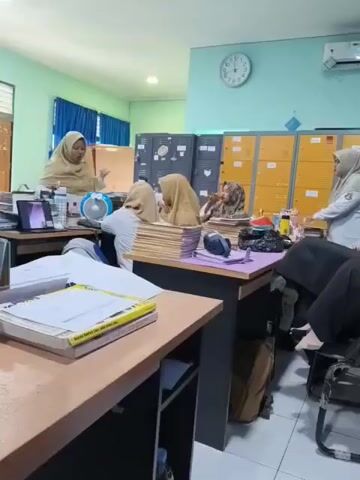 The video shows an indoor scene that appears to be an office or classroom setting. Several individuals, all wearing hijabs, are visible. One person in the foreground is sitting at a desk and looking towards the camera, with a laptop open in front of them. To the left of this individual, another person is seated at a desk with a fan and papers. In the middle ground, two individuals are seated at a table piled high with documents, with one of them appearing to be looking down at something. To the right, a person is seated at a desk, looking away from the camera. The room has a light blue wall and is furnished with desks, chairs, filing cabinets, and lockers. A clock is visible on the wall. The overall atmosphere suggests a work or study environment.
