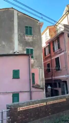 The video shows a town square in Italy, surrounded by colorful buildings. The buildings are painted in various shades of pink, yellow, and green. Some have green shutters on the windows. The square is paved with grey stones and there are some tables and chairs set up, presumably for outdoor dining. A few people can be seen walking around the square. There is a sign in the foreground that says "Vietato oltrepassare in linea gialla. Do not go beyond the yellow line." The sky is blue and clear.