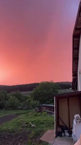 The video captures a slow panoramic view of a rural landscape under a dramatically colored sky. The camera pans from right to left, revealing a vast expanse of sky dominated by intense shades of red, orange, and hints of purple, suggesting either a vivid sunrise or sunset. Clouds are visible, illuminated by the sun to create these striking hues.

In the foreground and mid-ground, there is a garden area with visible tilled earth and green vegetation. Several small, covered structures, resembling greenhouses, are situated within the garden. A white, single-story house with a dark roof, a white door, and a window with closed blinds, featuring a satellite dish on its roof, becomes visible on the left side of the frame. On the far right, a portion of a wooden structure is visible. The background consists of rolling hills covered with dense forests. The scene depicts a peaceful, country setting. Human conversation can be heard in the background, but no people are visible in the frame. There is no visible text. The setting is consistent with a rural area in Kosten, Bulgaria.