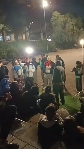 A group of approximately 20 people are gathered outdoors at night. Most of the people are standing in a circle on a small patch of artificial turf, while others are seated on concrete steps. A man in a white jacket is speaking and gesturing with his right hand. There is a street light and at least one portable light illuminating the scene. Some people are holding up their phones to record. One person is holding a camera. The ground is sandy. There are trees and buildings visible in the background. It appears they are shooting a video of the man.
