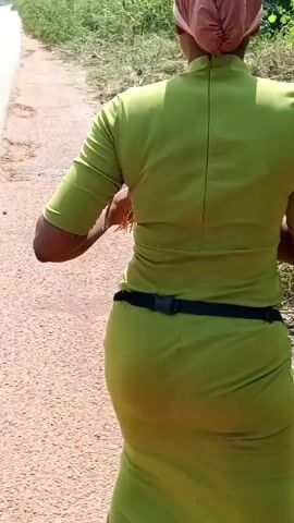 A woman in a long, olive-green dress and a black belt is walking on a dirt path through some greenery. She has a brown head covering and appears to be gathering something from the plants on the side of the path. Other people are visible in the background. The focus is primarily on the woman's body and her movements.