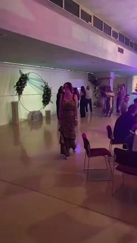 The video shows an indoor event, likely a party or reception, with people mingling and seated at tables. In the foreground, a woman in a patterned dress walks across the dance floor. To her right, chairs are arranged around a table. In the background, there are several round tables with white tablecloths, set with bottles, glasses, and candles. Some guests are seated at these tables, while others are standing and talking. A circular floral decoration is visible on the left side of the room. Lighting is dim and colorful, with stage lights and ceiling fixtures illuminating the space. The overall atmosphere appears festive.