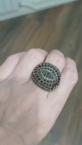A person's hand, adorned with a large, ornate ring, is shown in close-up. The ring features a silver, cage-like design surrounding a central eye-shaped pattern filled with small, dark stones, possibly black diamonds or cubic zirconia. The background is blurred, but appears to be an indoor setting with wooden flooring and a hint of a wall or furniture. The lighting is soft, highlighting the texture of the skin and the details of the ring. The hand is positioned to showcase the ring, with fingers slightly bent.