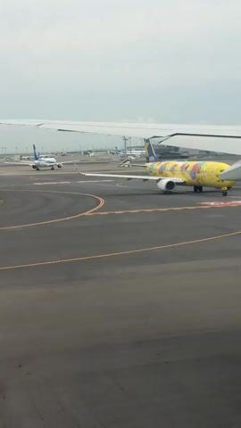 This is a low-angle, eye-level shot taken from inside an airplane. The camera is looking forward and slightly to the right, capturing the tarmac of an airport. A bright yellow airplane with colorful cartoon characters painted on its side is in the foreground, facing the camera. It is on a runway with orange and white markings. To the left of the yellow plane, another airplane, this one white with blue accents, is further down the runway and moving away from the camera. In the background, several other airplanes are parked at gates and terminals, with a control tower visible in the distance. The sky is overcast, with a light gray cloud cover. The video is shot from the perspective of someone in a window seat, as a portion of the wing of the current airplane is visible in the upper right corner of the frame. The yellow airplane appears to be stationary or slowly taxiing.