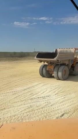 The video shows a side-view of a large, orange and tan dump truck driving across a dusty, light-colored terrain under a bright blue sky with scattered white clouds. The truck has four large, thick tires, two on each side of its rear axle. The bed of the truck is tilted upwards, and appears to be empty. The ground is flat and appears to be a construction site or quarry, with visible tire tracks in the light-colored dirt. The video appears to be filmed from the perspective of another vehicle.