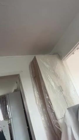 The video pans across a room. In the foreground, the red floor is visible. To the left, a white refrigerator is partially seen through a doorway. In the center and right of the frame, a large item, possibly a piece of furniture, is covered in clear plastic wrap. The walls are white, and the ceiling is also white with some texture. A window is visible in the upper right corner. The lighting appears to be natural light coming from the window.
