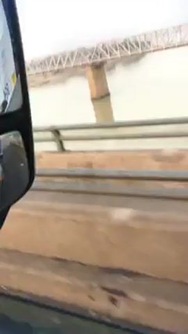The video shows a low-angle view of a metal bridge extending over water. The bridge appears to have a truss structure and is supported by brick pillars. The water is murky and grey, reflecting the overcast sky. A guardrail is visible in the foreground, suggesting the video was taken from another bridge or an elevated position. The camera seems to be moving, indicating a vehicle or person passing along the lower bridge.