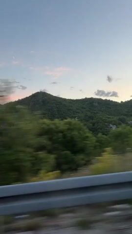 The video, shot from a moving vehicle, depicts a landscape of rolling, verdant hills against a dusk sky tinged with pink. A grey guardrail is visible along the bottom edge of the frame, indicating the perspective is from a roadway. The hills are densely covered with trees, creating a blanket of green broken by occasional rocky outcroppings. The sky is a gradient of light blue fading into pink hues near the horizon, dotted with scattered clouds. The camera is moving at a steady pace, giving a sense of traveling through the terrain.