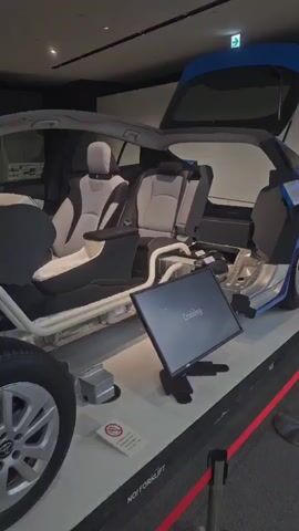 The video shows a cutaway display of a blue Toyota Prius car, with its hood and trunk open, revealing its internal components. The seats are visible inside the car, along with various mechanical parts in the engine bay and under the car. A monitor displaying the word "Cruising" is positioned in front of the car. The display is roped off with red tape, suggesting it is part of an exhibition or museum. The lighting is artificial, typical of an indoor environment.