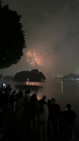 A low-angle, wide shot shows a fireworks display over a body of water at night, observed by a crowd of people standing on the shore. Silhouetted figures form the foreground, their shapes discernible but lacking specific details. A dark tree, partially visible on the left side of the frame, frames the scene. Across the water, a tree-covered islet stands out, its image reflected on the calm surface of the water. Fireworks burst in the sky, creating colorful explosions of red, white, and green. The reflection of the fireworks adds to the spectacle, creating a visually dynamic scene.  The overall tone is celebratory, despite the low lighting conditions.