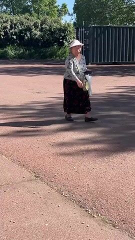 A middle-aged woman with fair skin is standing on a paved, sunlit surface. She is wearing a light-colored long-sleeved shirt with a floral design, a long dark skirt with floral patterns, and black shoes. She has a light-colored sun hat with a visor on her head. She also has a light-colored shoulder bag, and she is holding a white plastic bag in her left hand and a smartphone in her right. Behind her is a large black metal shipping container, green trees, and shrubbery. The ground is reddish-brown and covered in small pebbles, and some of it is in shadow.