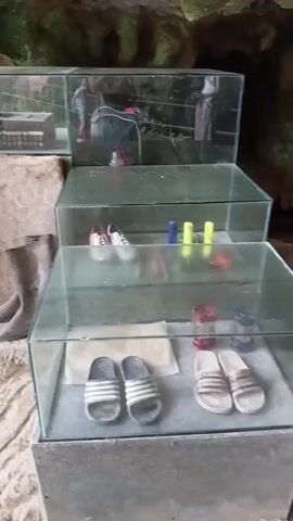 The video pans across a dimly lit, cavernous space. In the foreground, a pair of brown rubber boots rest on a textured, rocky surface. To the right, a multi-tiered glass display case holds various items. The top shelf contains what appears to be a collection of small objects, possibly tools or trinkets, along with a red and blue hard hat. The middle shelf displays a pair of pink and white sneakers and several colorful objects, some appearing to be markers. The bottom shelf holds a pair of black and white striped slides and two pairs of white slides with a similar stripe pattern. Behind the display case, a large burlap sack leans against the wall, and a coiled black cable lies on the ground. Further back, another glass-fronted display case is visible, with more items on its shelves. A small generator-like machine with an orange frame sits on the floor to the left of the display cases, next to some electrical cords. The walls of the space are rough and natural, suggesting a cave or an unfinished structure. The overall impression is of a storage area or an exhibit, possibly related to exploration or work in a rough environment.