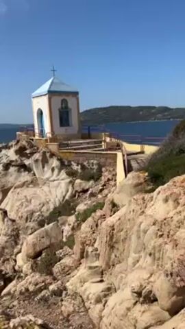 The video shows a small white and blue chapel perched atop a rocky cliff overlooking a body of water. The chapel has a cross on its roof and a blue-framed window. A short staircase and a metal railing lead up to it. The surrounding rocks are light tan and scattered with patches of green vegetation. In the background, across the water, is land with sparse trees. The sky is clear blue with a few wispy clouds. The camera is mostly still, providing a steady view of the scene.