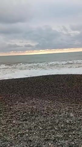 The video shows a pebble beach on a cloudy day.  The waves are crashing onto the shore.  The sky is overcast with a hint of light on the horizon.  There are no people visible in the video.