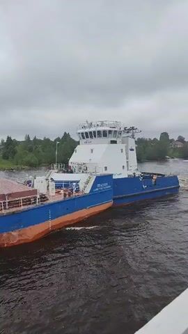 A mid-shot captures a blue and white boat traveling down a river. The boat is labeled "PT B2005," "САНКТ-ПЕТЕРБУРГ" (St. Petersburg), and "АЛЬТАИР" (Altair). The boat appears to be a tugboat connected to a barge. The river is choppy, and trees line the shore in the background. The sky is overcast and grey.