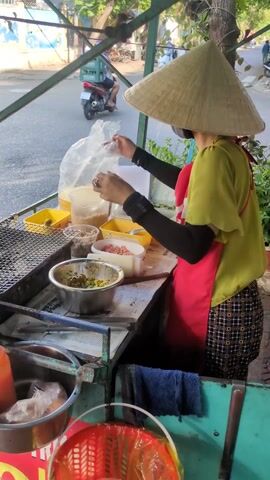 A street vendor in Vietnam prepares food at a mobile cart. The vendor, wearing a traditional conical hat, a green shirt, a red apron, and a face mask, arranges ingredients on the cart's surface. Various containers holding different food items are neatly organized. A grill sits to the side, with round wrappers placed on it. The cart is parked on a sidewalk next to a street, with traffic passing by. The environment includes trees and buildings in the background, providing context to the street scene.