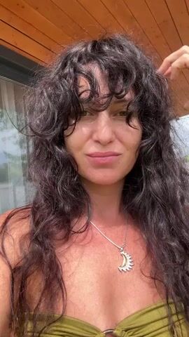 A woman with long, dark, curly hair and bangs is shown. She is wearing a green bikini top and a silver necklace with a sun and moon pendant. She is outdoors on a wooden deck. She smiles and touches her hair and eye in different clips.