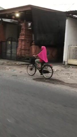 A person wearing a pink hijab and a patterned outfit is riding a bicycle. They are moving from left to right across the frame. The bicycle is black with a basket on the front. The person's feet are visible, wearing sandals.

The setting appears to be a street in an urban or semi-urban area, possibly in Southeast Asia, given the architectural style. Behind the cyclist, there is a building with intricate brickwork, reminiscent of traditional Indonesian architecture. A black tarp or fabric hangs over part of the building. Another building with light blue walls is visible further in the background. The ground is unpaved and dusty.

The activity is simply riding a bicycle. The time of day appears to be late afternoon or early evening, with soft lighting. The weather seems to be fair, with no visible rain or strong winds. No text is visible in the media file. The overall mood is casual and everyday.