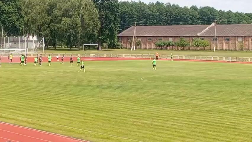 The video shows a soccer game taking place on a large, well-maintained grass field. A group of players, mostly wearing green jerseys and black shorts, are clustered on the left side of the field, near a goalpost. Another player in a green jersey is running towards the right side of the field, away from the group. There is a second goalpost visible in the background on the left, further up the field. On the right side of the field, there are two players wearing orange tops and black shorts. A few other players are scattered around the field. In the background, there is a red running track surrounding the field, and beyond that, a line of trees. Behind the trees, there is a long, brick building with a brown roof. The sky is overcast. The camera appears to be positioned high up, possibly in a stadium seating area.