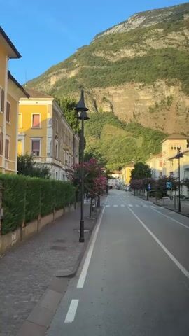 The video captures a view from inside a moving vehicle, looking out at a street scene in a town nestled at the base of a large, rocky mountain. The buildings on the left are primarily yellow, with white trim and brown shutters on the windows. A tall, dark lamppost stands prominently on the sidewalk. There are neatly trimmed green hedges lining the sidewalk. Several trees with bright pink flowers are visible along the street, adding color to the scene. The street itself is paved and appears to be a main road, with clear lane markings. The mountain dominates the background, its green, vegetated slopes rising steeply. The sky is clear and blue, indicating fair weather. The time of day appears to be late afternoon or early evening, given the warm light.