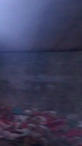 The video shows a blurred view from a moving vehicle, likely a train. Initially, a concrete wall covered in graffiti is visible. The camera then pans to show a rocky terrain with sparse vegetation and distant buildings with lights. The overall environment appears to be outdoors, possibly near a highway or railway track. The lighting is dim, suggesting dusk or night.