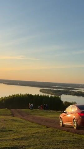 The video shows a scenic view of a river at sunset. The sky is a mix of blue and orange hues. A group of people is standing on a grassy hill overlooking the river. A car is parked nearby. The sun reflects brightly on the water. Two people walk along a dirt path on the hill.