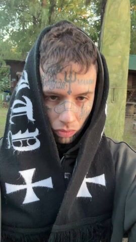 A man is recording a selfie video as he walks along a wooden deck towards a body of water. The man has numerous tattoos on his face and neck and a bandage on his nose. He is wearing a dark hoodie and a black scarf with white letters and crosses printed on it, which is pulled up over his head. He is looking at the camera, then off to the side and then back to the camera. The sun is setting behind him over a body of water with small boats and buildings in the background. The background includes trees, grass, and other buildings.
