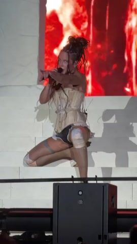 A woman with dark, messy hair is performing on stage. She is wearing a light-colored corset with straps, black underwear, and thigh-high stockings with bandages or wraps around her knees. She is holding a microphone in her right hand and her left arm is bent at the elbow. She is in a crouching position, with her right leg bent and her left leg extended back. Behind her is a large screen displaying a blurry image of flames in red and orange. In the foreground, a black speaker is visible. The stage appears to be made of white blocks.