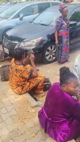 The video shows a group of women sitting on the ground next to a row of parked cars on a brick pathway. They appear to be working on documents, possibly filling out forms, with some holding books and using their phones. Other individuals, including a man with a backpack, are walking by. The cars are parked in a row and trees are visible in the background.