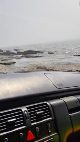 The video shows a view from inside a car looking out at a body of water. The car's dashboard and side door are visible in the foreground. Outside the car, the water appears calm with some rocks or land formations in it. The sky is overcast and hazy. There are no people visible in the video.