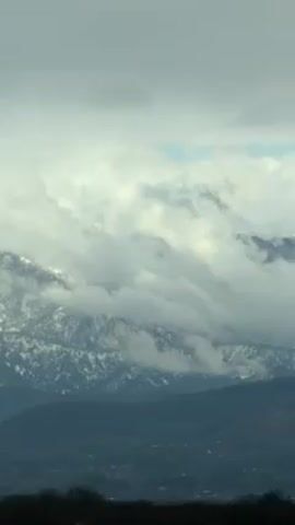 The video shows a mountain range partly covered in snow. The mountains are also partially obscured by clouds, and the sky above is overcast. The lower part of the frame shows a darker landscape with trees. The clouds move slightly throughout the video.