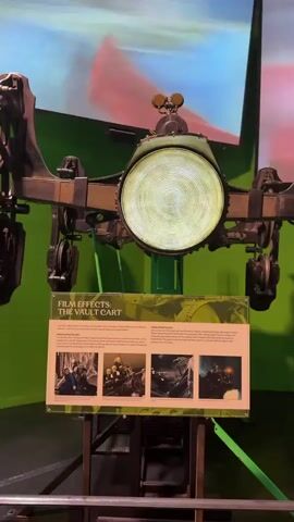 This media file captures a display related to the Harry Potter films, likely at Warner Bros. Studio Tour London – The Making of Harry Potter in Watford, United Kingdom. The central object is a "Vault Cart" prop, a steampunk-esque contraption with a large, illuminated circular lens and mechanical arms with wheels. It's mounted on a stand.

In front of the cart is an informational display board. The board's title reads "FILM EFFECTS: THE VAULT CART". It features text describing the cart's use in the films and four smaller images showcasing scenes and behind-the-scenes shots, likely from the Harry Potter movies, one of which appears to be from Harry Potter and the Philosopher's Stone.

Behind the cart, a large screen displays a clip from a Harry Potter film, featuring a close-up of Daniel Radcliffe as Harry Potter with an intense expression. The background of the display is a vibrant green wall.

In the lower section of the frame, several people are visible, their backs turned to the camera, observing the exhibit. There are also two motorcycles with sidecars on display, possibly props from the films as well. The overall setting appears to be an indoor exhibition space, indicated by the lighting and the presentation of the artifacts. The time of day is indeterminate.