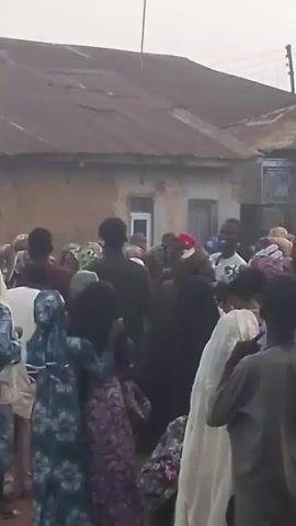 A crowd of people, many wearing headscarves and traditional clothing, are gathered outdoors in front of a building with a corrugated metal roof. Some individuals are interacting with each other, with one person appearing to be speaking to the group and others gesturing. The atmosphere seems lively, possibly a celebration or a community event. The background shows a simple building with windows and a rough, neutral-colored wall. There are no visible vehicles or signs of commercial activity. The lighting suggests it is daytime, possibly overcast.