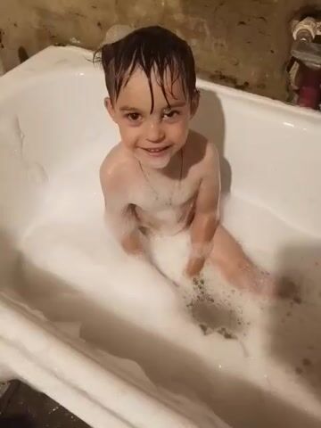 A child is sitting in a bathtub filled with foam. The child is looking at the camera and smiling. The child then turns their head and covers their face with their hands. The child then looks back at the camera and smiles again. The bathtub is white and appears to be in a bathroom. The walls of the bathroom are a dark color. There is a bottle of shampoo on the edge of the bathtub.