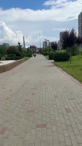 This video shows a paved walkway through a park on a sunny day with scattered clouds. The walkway is made of light grey paving stones with interspersed red stones. On either side of the walkway are well-maintained green lawns, decorative bushes, and trees. In the background, several tall buildings can be seen, suggesting an urban or suburban setting. A person is walking away from the camera on the walkway in the distance. Further down the path, another person can be seen sitting on a bench, possibly taking a break or enjoying the scenery. A large, fluffy white cloud is prominent in the bright blue sky, indicating fair weather. The scene appears peaceful and serene, with the natural elements of the park contrasting with the urban landscape in the background. The time of day appears to be daytime, likely mid-morning or afternoon, given the bright sunlight. No visible text is present in the video. The location context is Krasnodarskiy, Russia.