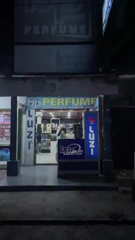 The video shows the exterior of a perfume shop at night. The shop's sign reads "BP PERFUME" in large yellow letters above the storefront. There are also vertical signs on either side of the entrance that say "LUZI". Inside the shop, a person is visible behind a counter, arranging products. The shop appears to be well-lit, with various bottles and displays of perfume visible. The street in front of the shop is dark.