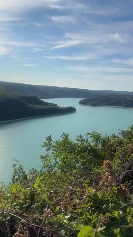 The video captures a panoramic view of a vast, turquoise lake surrounded by lush, green, tree-covered hills under a bright blue sky with scattered white clouds. The water appears calm, with a few small boats visible on its surface. In the foreground, there is a dense cluster of green foliage, including leaves and branches, partially obscuring the view of the lake and hills. The lighting suggests it is daytime, likely late morning or afternoon, given the clear visibility and the shadows cast. The overall scene depicts a natural landscape, possibly a scenic overlook of a reservoir or a large natural lake.