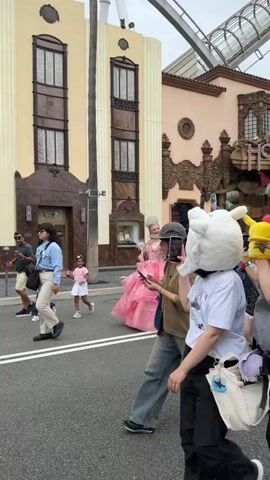 A daytime video captures a lively street scene in what appears to be a theme park in Osaka, Japan. A diverse group of people, including adults and children, are walking along a paved road flanked by multi-story buildings.

Several individuals are dressed in elaborate costumes: one person wears a large white fluffy animal head, another a large yellow fluffy animal head (resembling Pikachu), a person is dressed as a witch with green skin, a black cloak, and holds a broomstick, and another is in a vibrant pink princess gown. These costumed characters appear to be part of an event, with onlookers observing and photographing them.

Other people are dressed in casual attire; some carry bags or backpacks, and one individual has a baby carrier. Many people are seen holding smartphones, actively capturing the moment.

The architecture of the buildings in the background suggests an old Hollywood or European style. One prominent building features a sign that reads "MAX FACTORY STUDIO" and the number "6277" below it. In the upper background, structures resembling a roller coaster track are visible. The weather appears to be overcast or partly cloudy, and the overall atmosphere is busy and engaging.