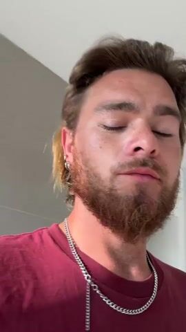 A man with blonde hair, a beard, and earrings is wearing a maroon t-shirt and a silver chain. He is looking at the camera and moving his head. The background appears to be a room with light-colored walls and a window.