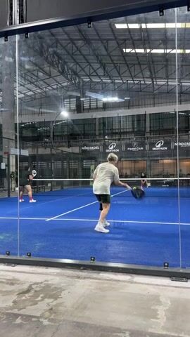 Two individuals are playing padel tennis on an indoor blue court surrounded by glass walls. The person in the foreground, wearing a light-colored t-shirt and dark shorts with a knee brace, is in the process of hitting a shot with a padel racket. Another person, wearing a black t-shirt and shorts, is visible on the other side of the net, also holding a racket. The court has white lines marking its boundaries. The background shows the ceiling structure of the indoor facility, with industrial lighting and several logos displayed on the back wall, including "NINO PADEL", "PRACTICE CLUB", and "DECATHLON". The floor outside the court appears to be a concrete surface.