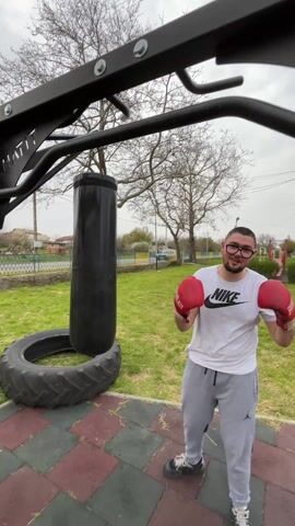 A man wearing a white t-shirt, grey sweatpants, glasses, and red boxing gloves stands outside on a red and grey tiled surface, with a grassy area and playground in the background. He is standing next to a large black punching bag hanging from a black metal frame, and a tire sits nearby. The man raises his gloved fists and appears to strike at the camera as it moves quickly forward.