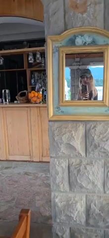 The video shows a woman taking a selfie in a mirror. The mirror is framed in gold and mounted on a stone wall. The woman is wearing a green baseball cap and a gray tank top. She is holding a gray phone in her right hand and showing off a tattoo on her left arm. Through the mirror, we can see a view of a lake and another person sitting at a table. The background includes a wooden cabinet with oranges, glasses, and a kettle on it. The floor is paved with brick. The woman smiles and looks directly into the camera.