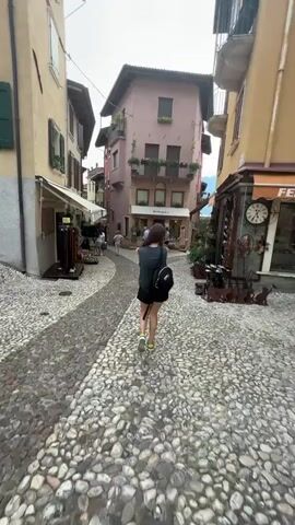 A young person wearing a black tank top, black shorts, and yellow sneakers walks away from the camera down a narrow cobblestone street. The person is carrying a black backpack. The street is lined with buildings, some of which appear to be shops. One building in the center is painted pink and has balconies with potted plants. The sky is overcast.