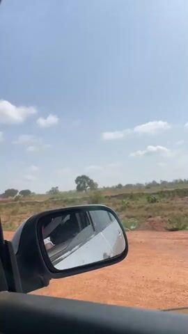 The video shows a view from a moving car, looking out the side window. The landscape consists of dry, scrubby vegetation and reddish-brown earth.  In the distance, there are scattered trees and the sky is light blue with some white clouds.  A car side mirror is visible in the foreground.