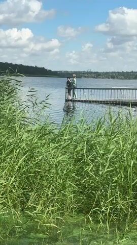 Two people stand on a pier by a lake. The person on the right, wearing a light green outfit, is touching the hair of the person on the left, who is wearing a black shirt and black leggings. The pier has a metal railing. Reeds are visible in the foreground. The sky is blue with white clouds.