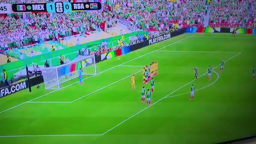 The video shows a soccer match between Mexico (MEX) and South Africa (RSA), with Mexico leading 1-0. The action is taking place near the goal, with players from both teams gathered. A group of players in yellow shirts are standing in a defensive wall in front of the goal. A player in a green and white uniform, presumably from Mexico, is preparing to take a free kick. Another player in green and white is in the penalty box, and the goalkeeper in a light blue uniform is in position. The stadium is filled with spectators, many of whom are wearing green and white, the colors of Mexico. The word "PLAY.FIFA.COM" is visible on a banner around the field.