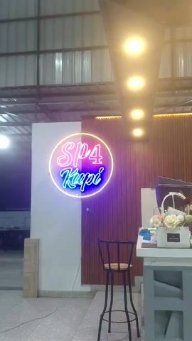 The camera pans across the interior of SP4 Kupi, a coffee shop, showing the front counter and the barista at work. The shop has a high, corrugated metal roof with pendant lighting.  A neon sign reading “SP4 Kupi” is fixed to the wall to the left of the counter. The counter is decorated with floral arrangements.  A young person in a black shirt and white pants stands to the right of the counter.