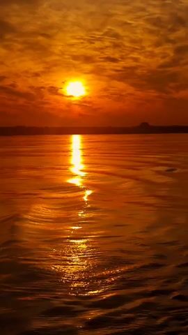 This video shows a wide shot of a body of water, likely a lake or ocean, during sunset. The sun, a bright orb of light, is partially obscured by wispy clouds, casting a warm, golden glow across the scene. The water's surface is textured with gentle waves, reflecting the sun's brilliance in a shimmering path that stretches towards the viewer. The overall impression is one of tranquility and natural beauty, with the colors of the sunset creating a dramatic and serene atmosphere.